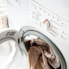Eine Waschmaschine mit offener Tür und Wäsche darin. Das Bedienfeld zeigt verschiedene Waschprogramme, aber die Wäsche riecht nach Kanal.