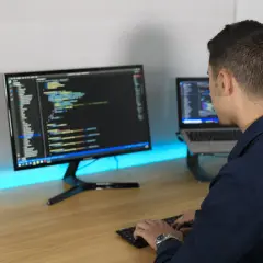 Ein Mann tippt Code auf einer Tastatur, während er auf zwei Bildschirme blickt. So macht ein Programmierer seine Arbeit.