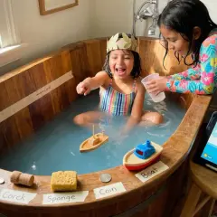 Zwei Mädchen spielen in einer Holzbadewanne, inspiriert von Archimedes' Entdeckung. Sie experimentieren mit Objekten wie Löffel, Korken, Schwamm und Münze, um zu sehen, was schwimmt.