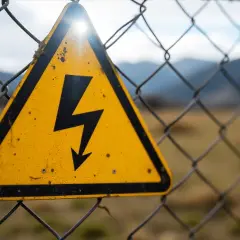 Gelbes Warnschild mit Blitzsymbol auf einem Maschendrahtzaun, das auf die Gefahren des elektrischen Stroms hinweist.