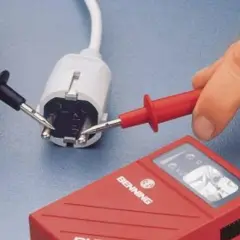 Hände mit Messspitzen an einem Stecker, um Strom zu messen. Ein rotes Messgerät liegt daneben.