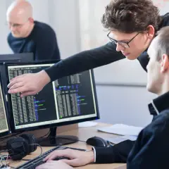 Zwei Programmierer am Schreibtisch, die auf Code auf zwei Monitoren zeigen. Was braucht man um Programmierer zu werden? Konzentration und Teamwork.