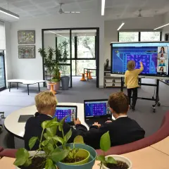 Kinder lernen an einer digitalen Tafel in der Schule. Ein Junge interagiert mit dem Touchscreen, während zwei Schüler an Laptops sitzen.