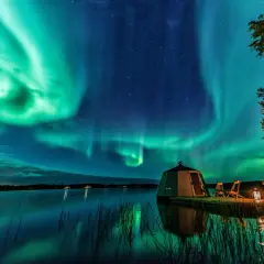 Grüne Polarlichter tanzen über einem See. Ein kleines Haus und zwei Stühle stehen am Ufer. So entstehen Polarlichter: geladene Teilchen der Sonne treffen auf die Erdatmosphäre.
