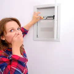 Schockiertes Gesicht einer Frau, die ihre Hand vor den Mund hält, während sie auf den Sicherungskasten zeigt. Eine Sicherung ist durchgebrannt.