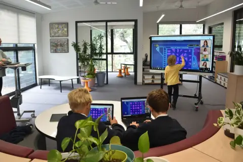Kinder lernen an einer digitalen Tafel in der Schule. Ein Junge interagiert mit dem Touchscreen, während zwei Schüler an Laptops sitzen.