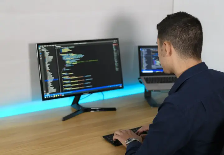 Ein Mann tippt Code auf einer Tastatur, während er auf zwei Bildschirme blickt. So macht ein Programmierer seine Arbeit.