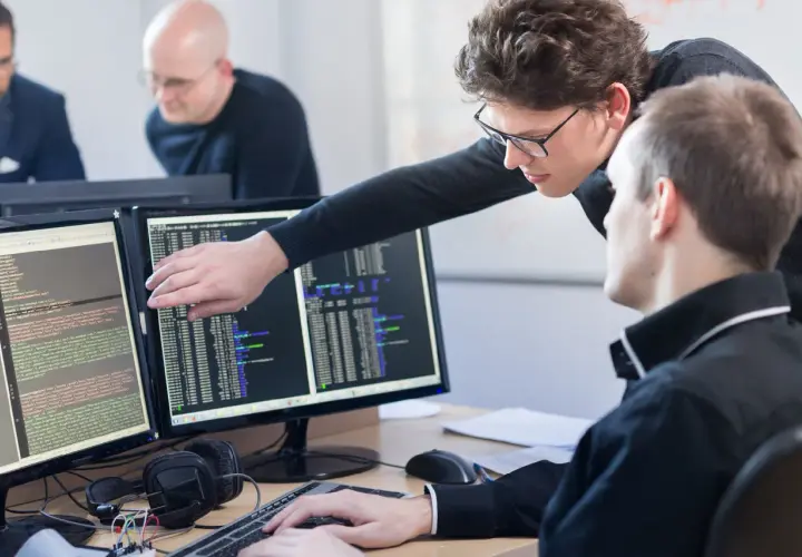 Zwei Programmierer am Schreibtisch, die auf Code auf zwei Monitoren zeigen. Was braucht man um Programmierer zu werden? Konzentration und Teamwork.