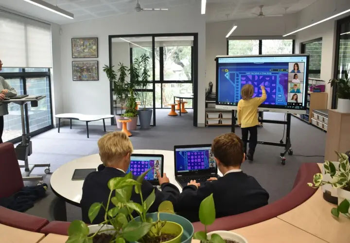 Kinder lernen an einer digitalen Tafel in der Schule. Ein Junge interagiert mit dem Touchscreen, während zwei Schüler an Laptops sitzen.