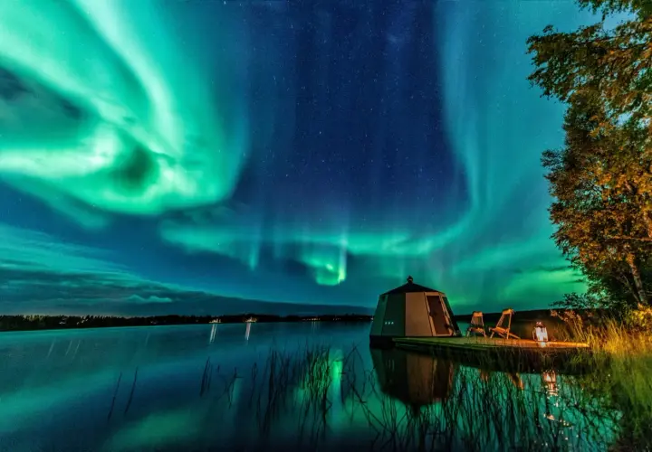 Grüne Polarlichter tanzen über einem See. Ein kleines Haus und zwei Stühle stehen am Ufer. So entstehen Polarlichter: geladene Teilchen der Sonne treffen auf die Erdatmosphäre.
