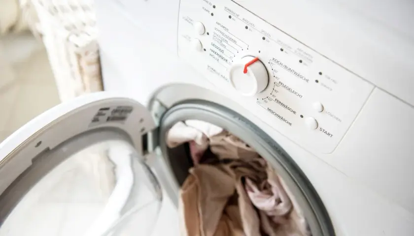 Eine Waschmaschine mit offener Tür und Wäsche darin. Das Bedienfeld zeigt verschiedene Waschprogramme, aber die Wäsche riecht nach Kanal.