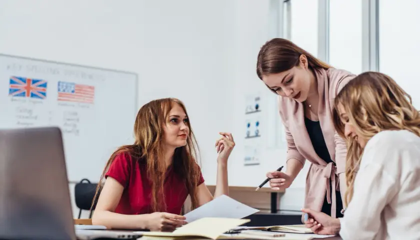 Drei Frauen lernen gemeinsam. Eine Lehrerin erklärt etwas, während die Schülerinnen aufmerksam zuhören. Auf dem Whiteboard sind die Flaggen Großbritanniens und der USA zu sehen, was auf den Unterricht in englischen computer sprachen hindeutet.