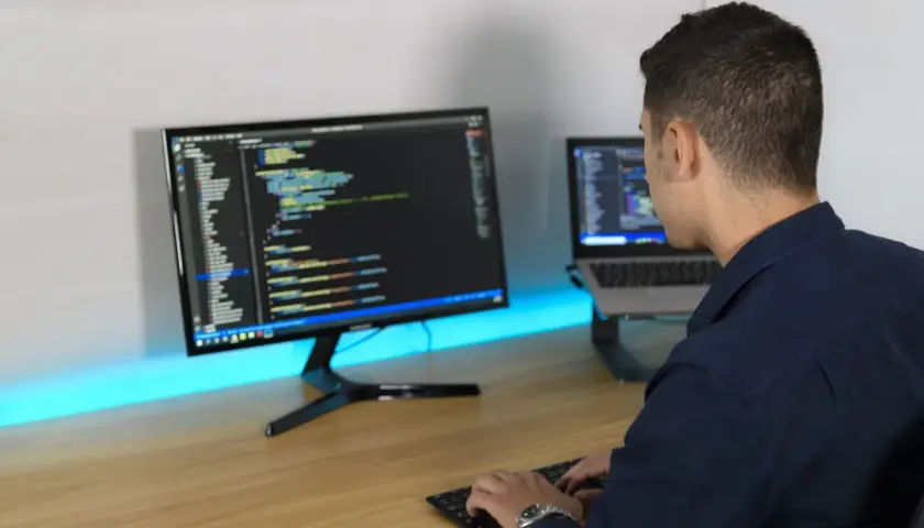 Ein Mann tippt Code auf einer Tastatur, während er auf zwei Bildschirme blickt. So macht ein Programmierer seine Arbeit.