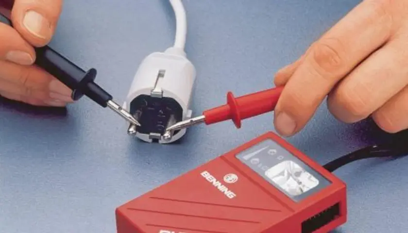 Hände mit Messspitzen an einem Stecker, um Strom zu messen. Ein rotes Messgerät liegt daneben.