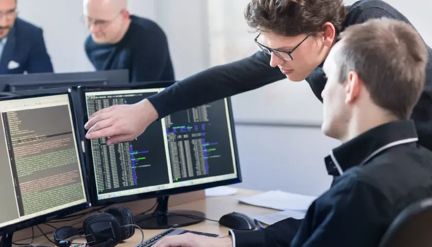 Zwei Programmierer am Schreibtisch, die auf Code auf zwei Monitoren zeigen. Was braucht man um Programmierer zu werden? Konzentration und Teamwork.