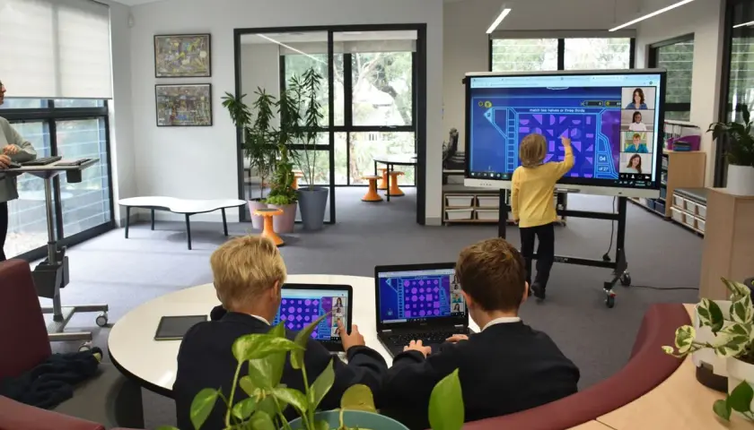 Kinder lernen an einer digitalen Tafel in der Schule. Ein Junge interagiert mit dem Touchscreen, während zwei Schüler an Laptops sitzen.