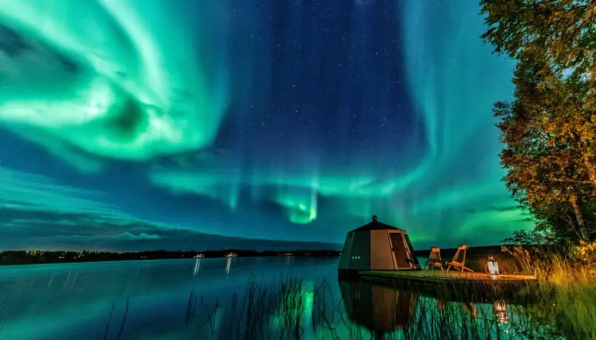 Grüne Polarlichter tanzen über einem See. Ein kleines Haus und zwei Stühle stehen am Ufer. So entstehen Polarlichter: geladene Teilchen der Sonne treffen auf die Erdatmosphäre.