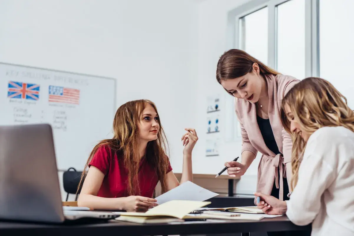 Drei Frauen lernen gemeinsam. Eine Lehrerin erklärt etwas, während die Schülerinnen aufmerksam zuhören. Auf dem Whiteboard sind die Flaggen Großbritanniens und der USA zu sehen, was auf den Unterricht in englischen computer sprachen hindeutet.