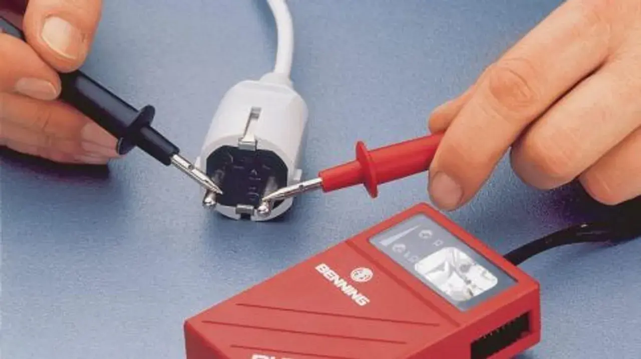 Hände mit Messspitzen an einem Stecker, um Strom zu messen. Ein rotes Messgerät liegt daneben.