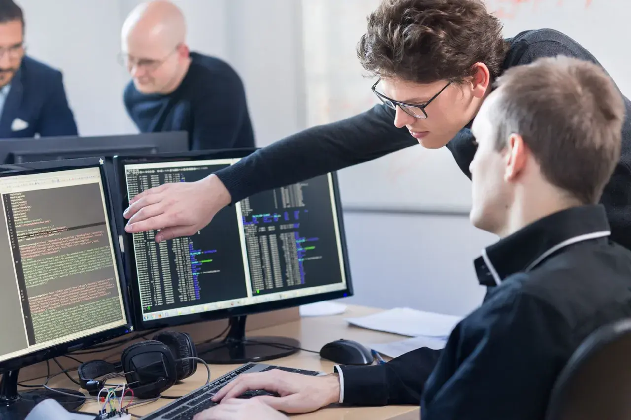 Zwei Programmierer am Schreibtisch, die auf Code auf zwei Monitoren zeigen. Was braucht man um Programmierer zu werden? Konzentration und Teamwork.