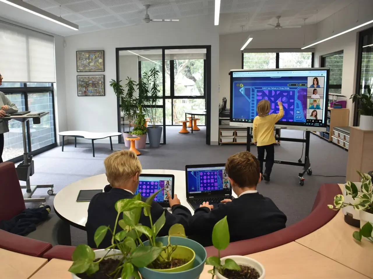 Kinder lernen an einer digitalen Tafel in der Schule. Ein Junge interagiert mit dem Touchscreen, während zwei Schüler an Laptops sitzen.