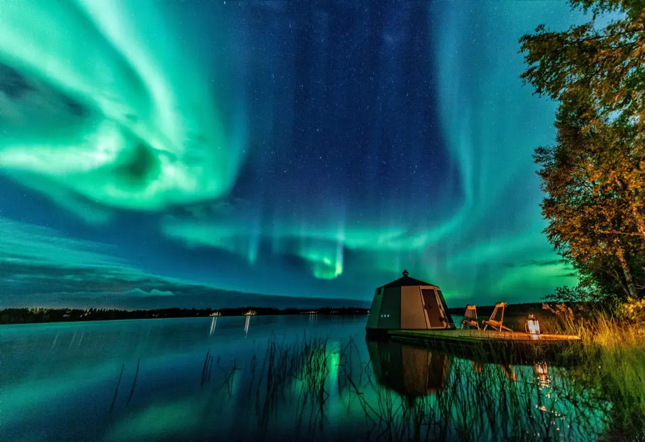Grüne Polarlichter tanzen über einem See. Ein kleines Haus und zwei Stühle stehen am Ufer. So entstehen Polarlichter: geladene Teilchen der Sonne treffen auf die Erdatmosphäre.